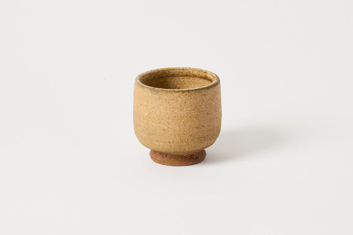 A small handmade ceramic cup with a textured, speckled beige and light brown glaze, standing on a small round base against a clean white background.
