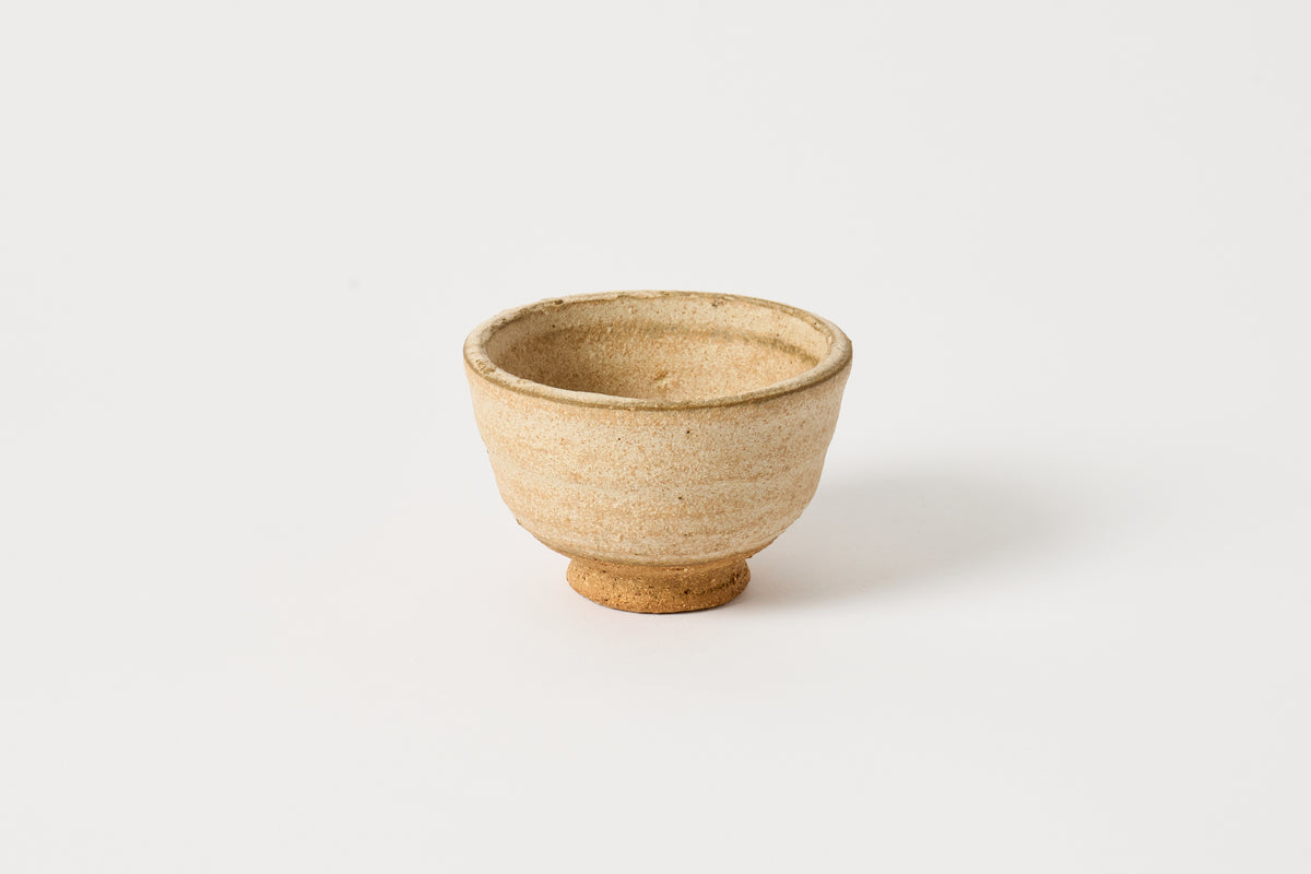 A small handmade ceramic tea bowl with a textured, speckled beige glaze and a natural clay foot, photographed on a clean white background.