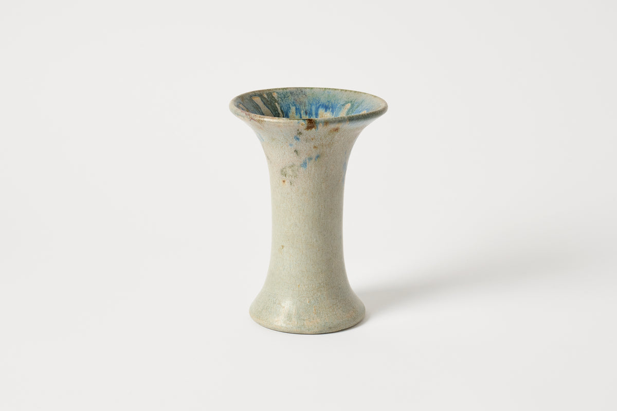 A small ceramic bud vase with a flared rim and a narrow cylindrical body, featuring a textured off-white glaze with blue and brown speckled accents inside and on the exterior, set against a plain light gray background.