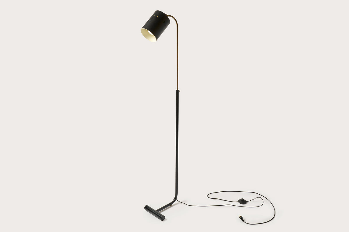 A floor lamp with a black cylindrical shade, a slim black metal stand, and a gold-finished central support rod, shown against a solid light beige background.