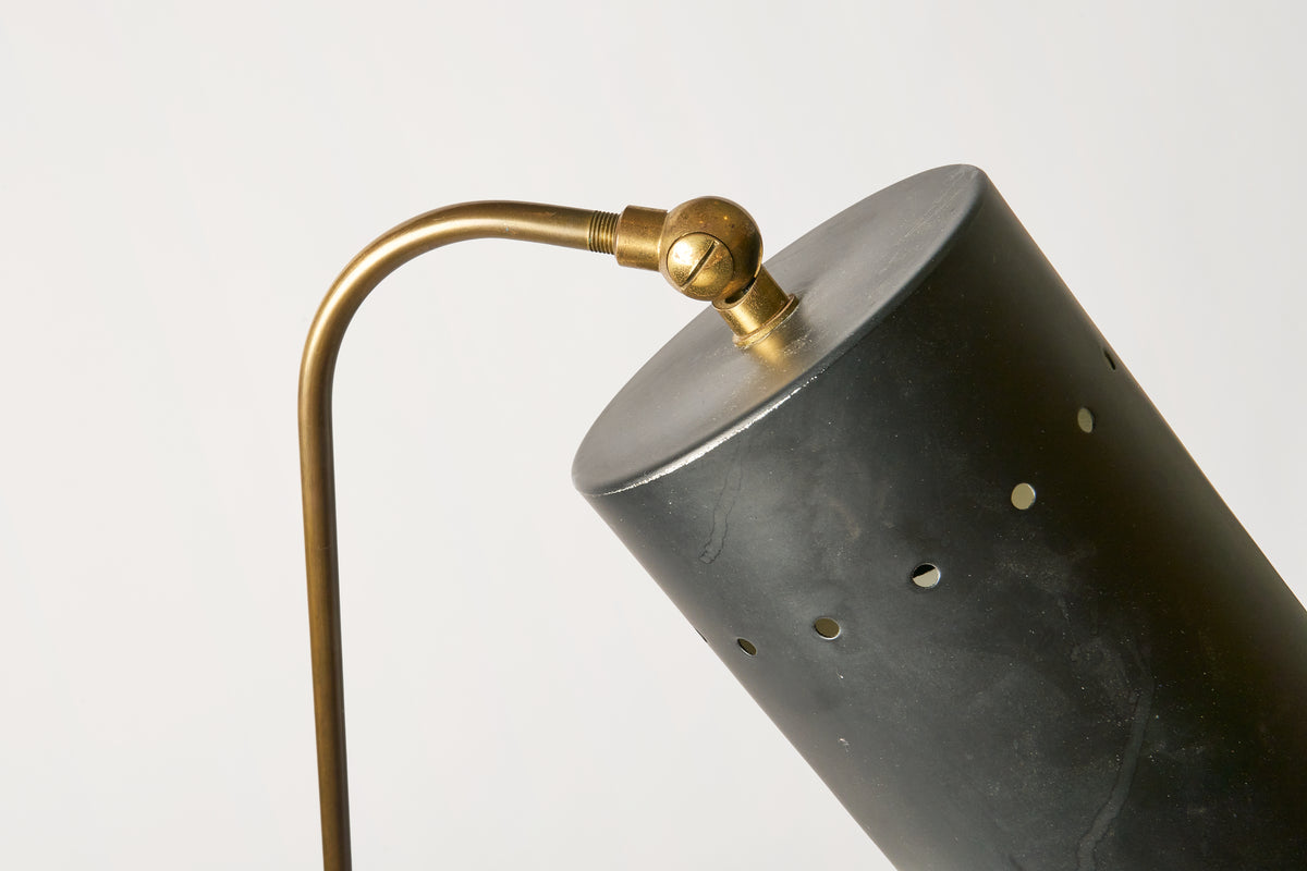 Close-up of a table lamp with a matte black cylindrical shade featuring small brass ventilation holes and a curved brass arm, set against a plain light gray background.