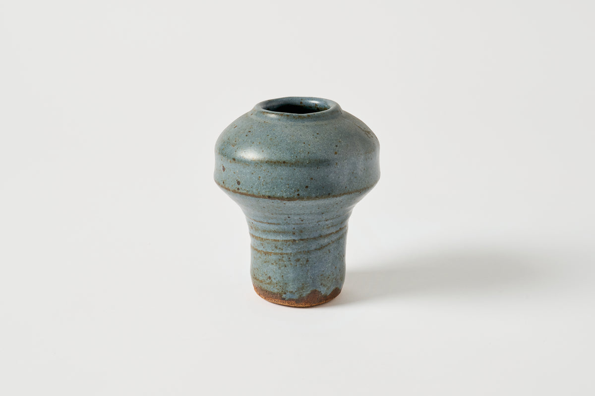 A small handmade ceramic vase with a rounded bulbous body and a narrow flared base, covered in a textured slate-blue glaze with subtle brown speckles and a natural terracotta edge, set against a plain light gray background.