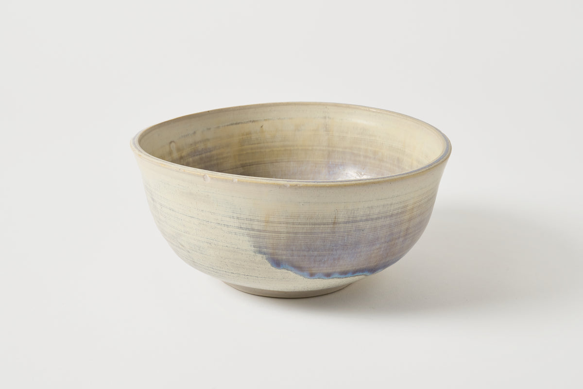 A handmade ceramic bowl with a textured, hand-painted glaze featuring a cream-beige exterior and a soft blue-purple interior, set against a plain light gray background.