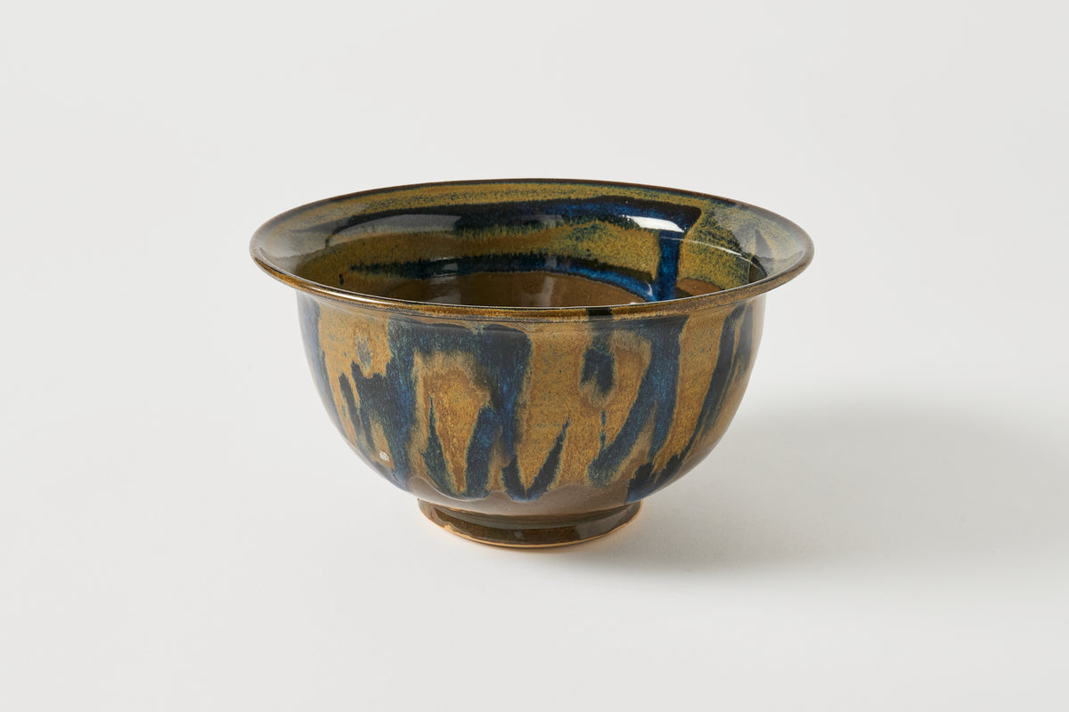 A handmade ceramic bowl with a glossy glaze featuring a textured gold and dark blue drip pattern, photographed on a clean white background.