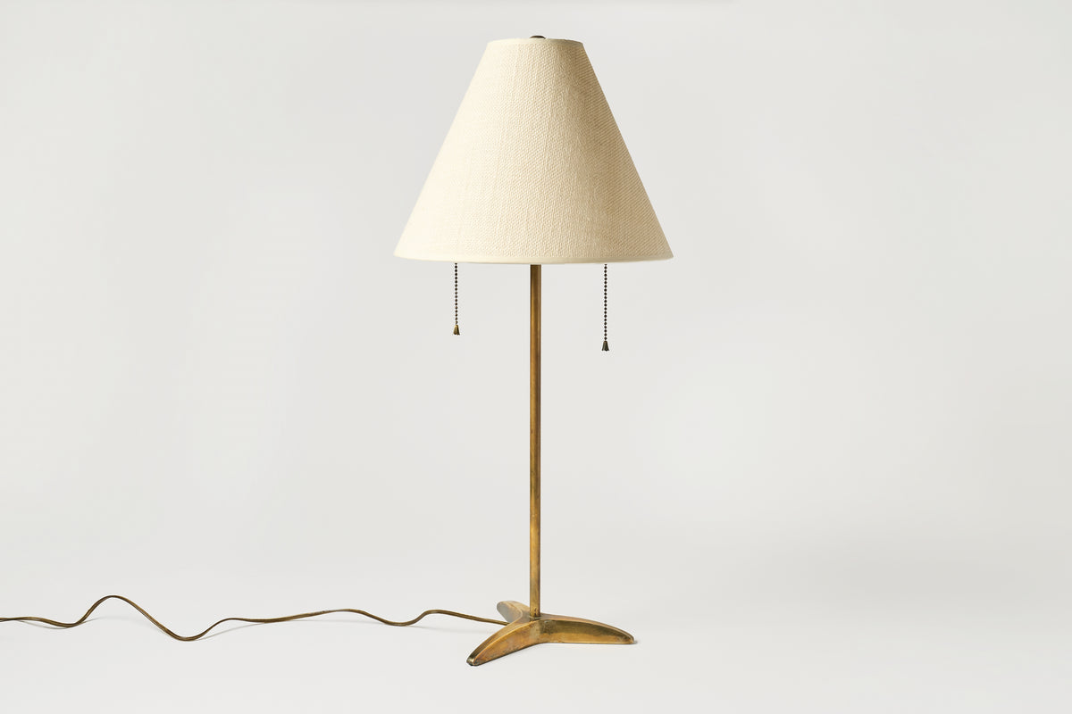A mid-century modern table lamp with a tapered cream fabric shade, a brass central stem, and a three-pronged brass base, shown against a plain light gray background with its cord visible.