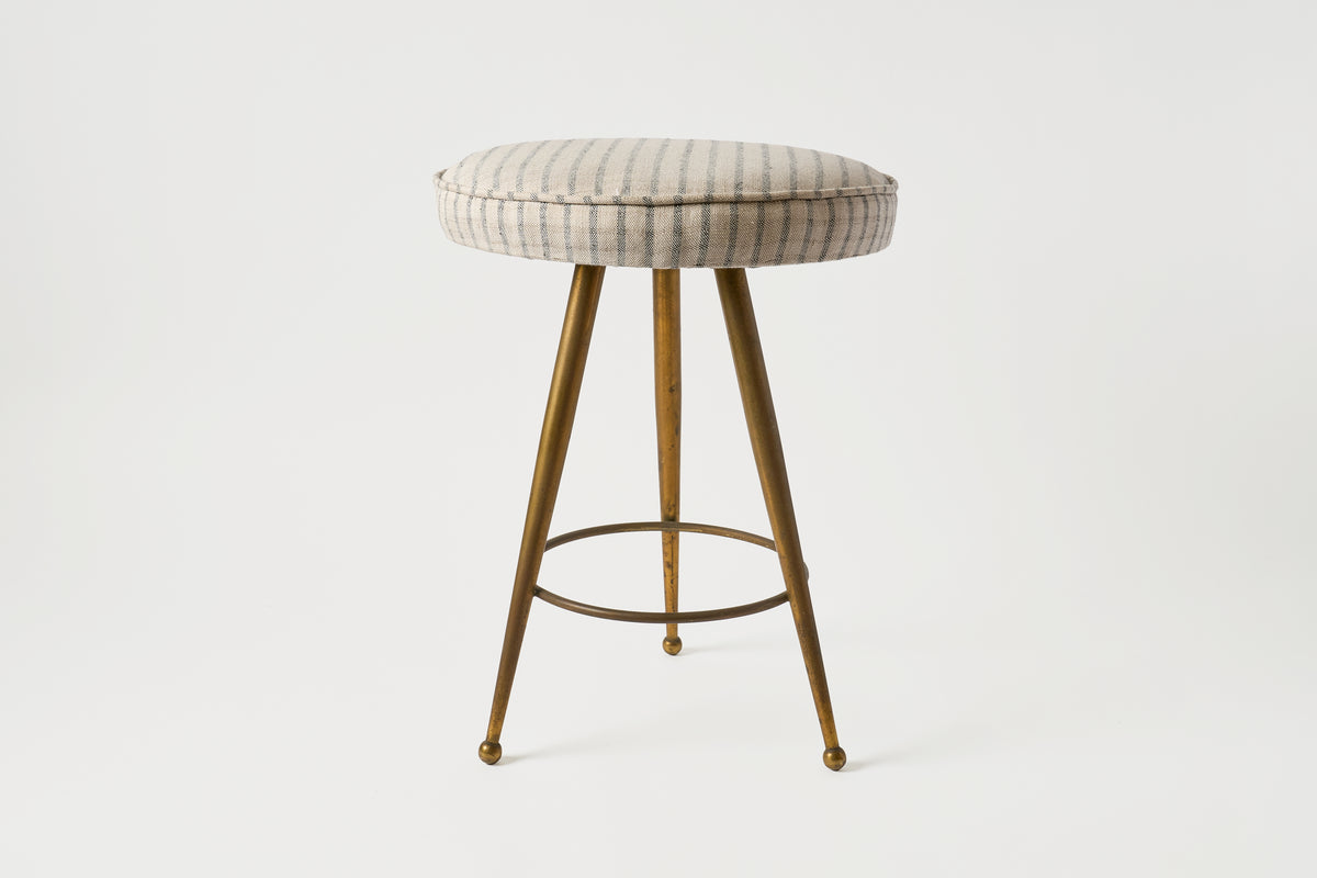 An upholstered bar stool with a round cushioned seat in a beige and gray striped fabric, supported by three tapered brass legs and a circular metal footrest, set against a plain white studio background.