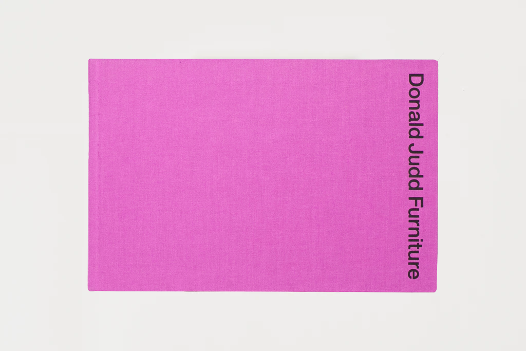 A bright pink rectangular fabric cover or book-style sleeve with black vertical text reading 'Donald Judd Furniture' on the right edge, shown on a light gray background.