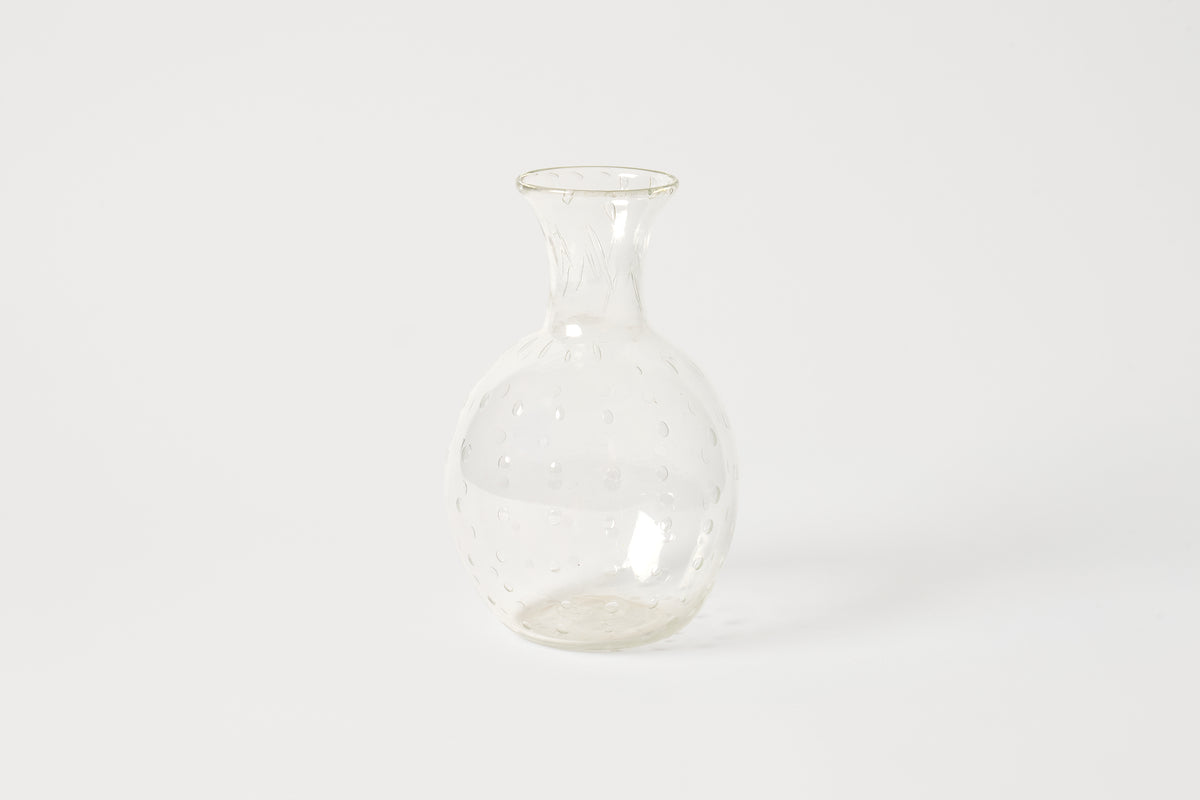 A small clear glass bottle with a bulbous body, narrow neck, and flared rim, featuring a subtle dotted texture, set against a plain white background.