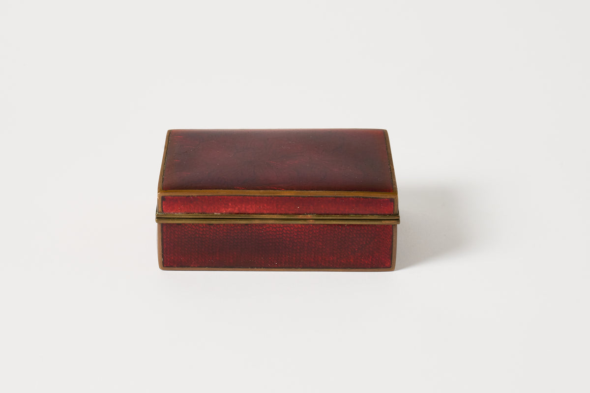 A small rectangular red leather or faux-leather box with gold-toned metal trim, photographed on a plain light gray background.