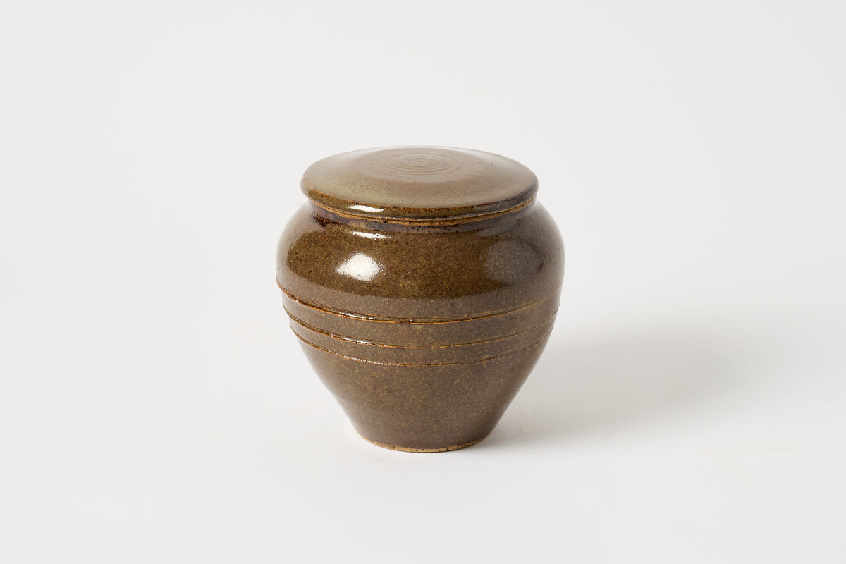 A small glazed ceramic lidded jar or urn in a speckled brown and olive-green glaze, photographed on a clean white background.