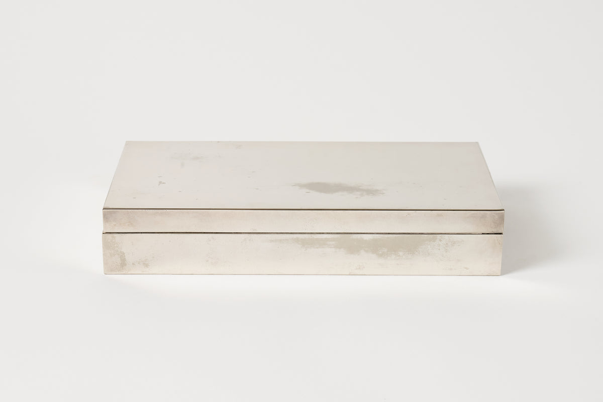A rectangular metal box with a hinged lid, featuring a brushed silver finish and subtle tonal variations, set against a plain white background.
