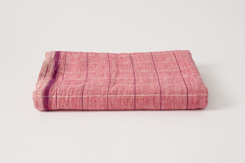 A folded pink woven textile, likely a blanket, throw, or scarf, featuring a subtle grid pattern and a dark purple stripe along one edge, set against a plain light gray background.