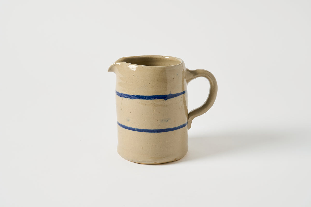A handmade ceramic pitcher with a beige stoneware body, featuring two horizontal blue painted stripes and a small pouring spout, set against a plain light gray background.