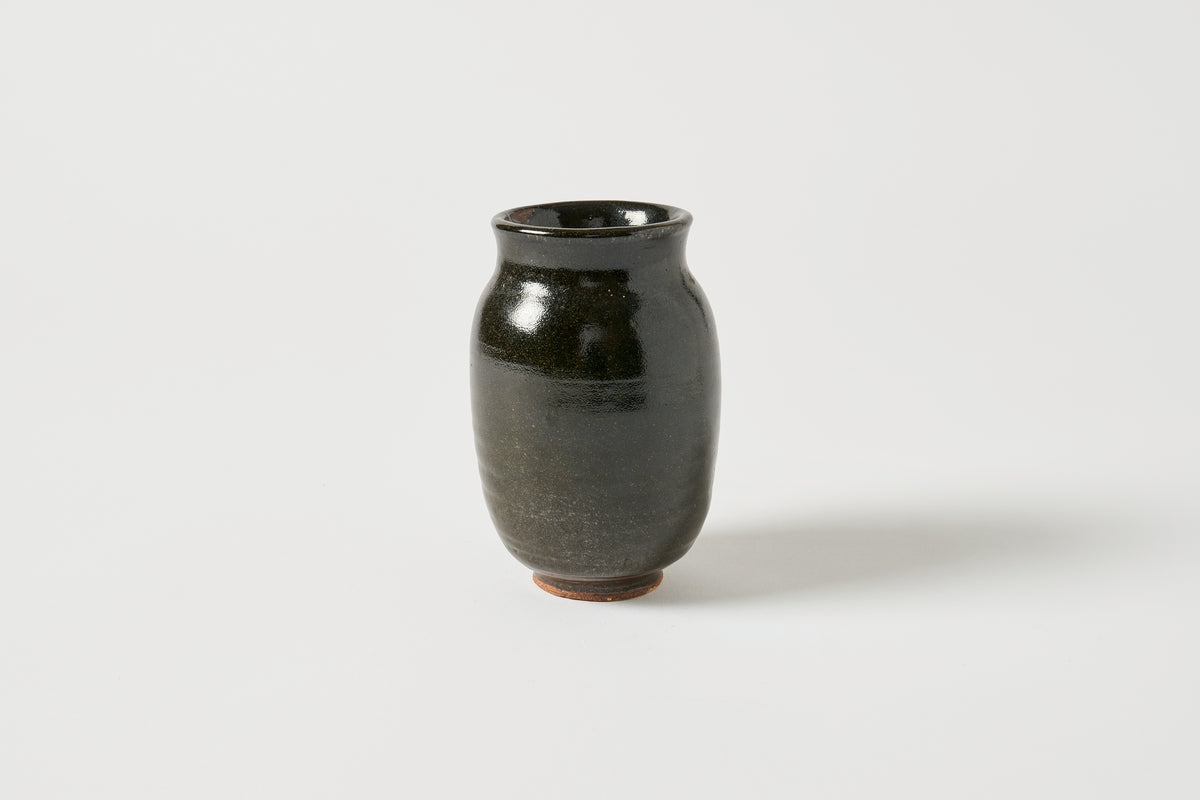 A small glossy ceramic vase with a dark green to black glaze and a subtle earthy brown base, photographed on a clean light gray background.