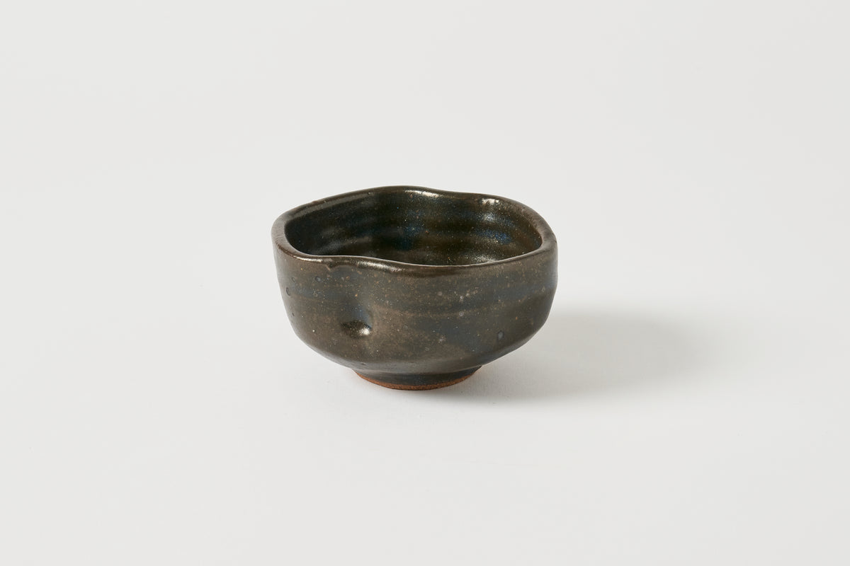 A small handmade ceramic bowl with a dark, rustic glaze and irregular organic shape, photographed on a clean light gray background.