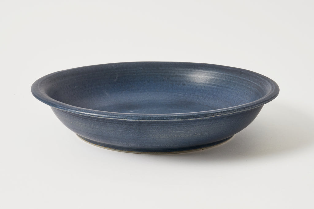 A round ceramic shallow bowl with a textured, matte dark blue glaze, photographed on a clean white background.