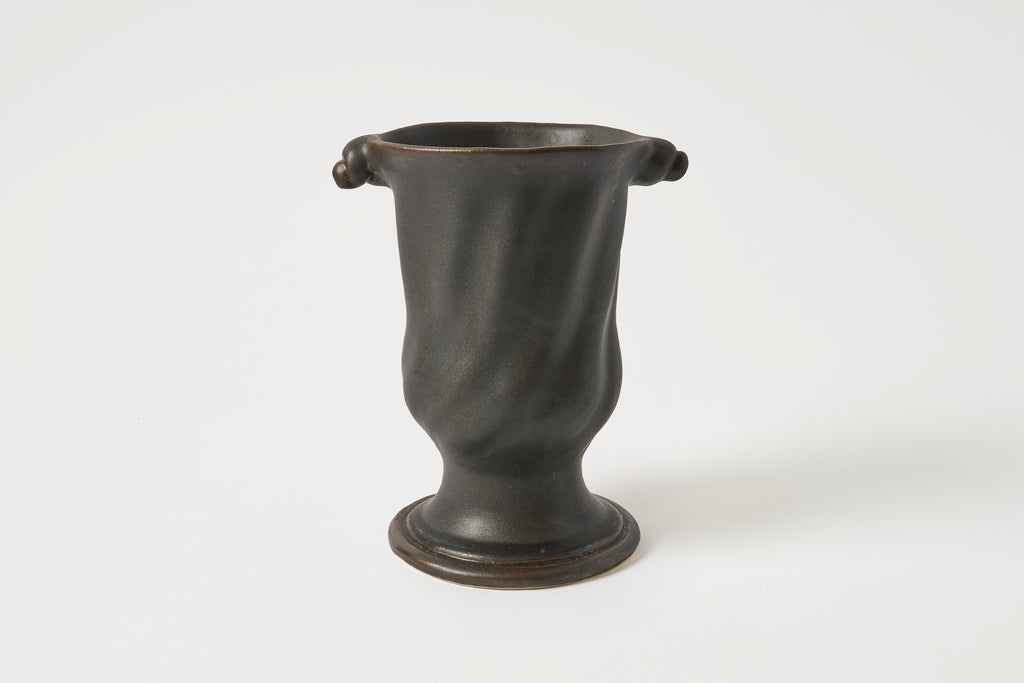 A dark, matte-finished ceramic vase with a flared base and two small loop handles near the rim, photographed against a plain light gray studio background.